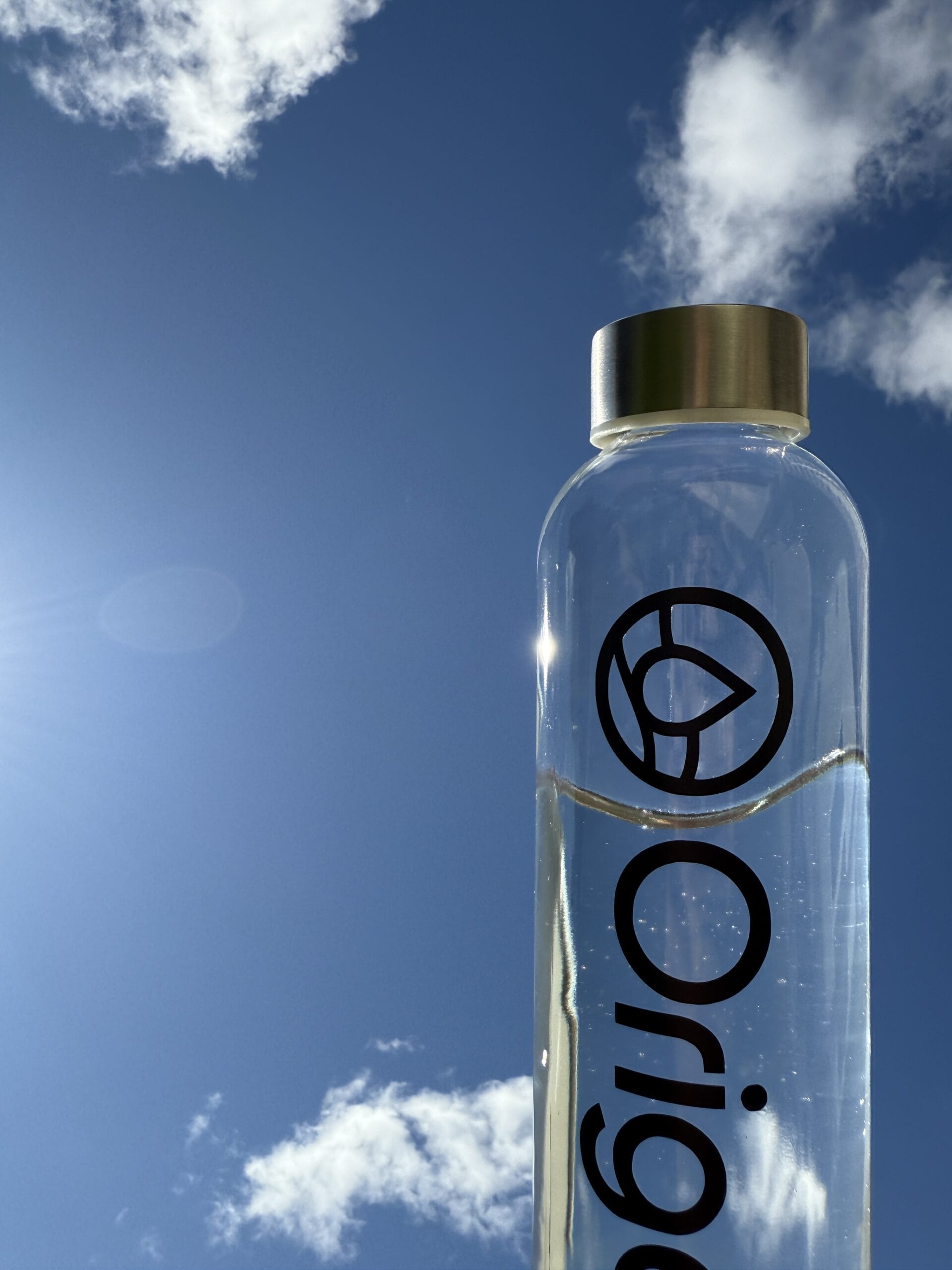 Clear water bottle with a metallic cap against a bright blue sky and fluffy white clouds.
