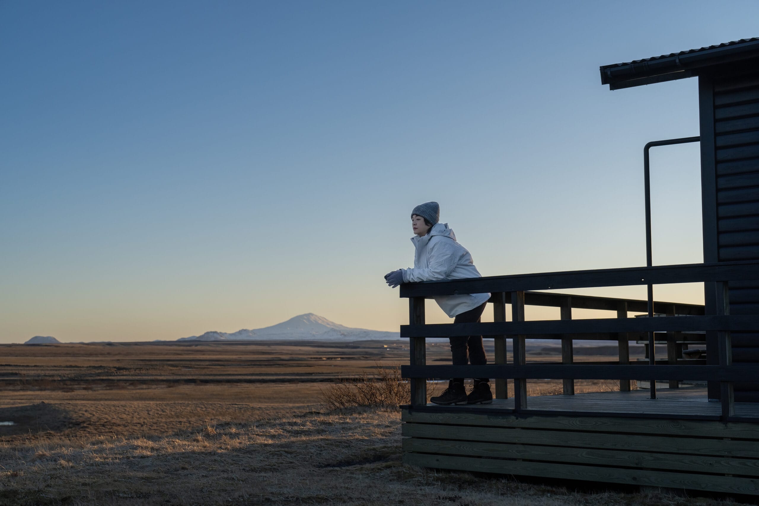woman leaning on log cabin porch terrace enjoying scenery view morning, mountains behind her