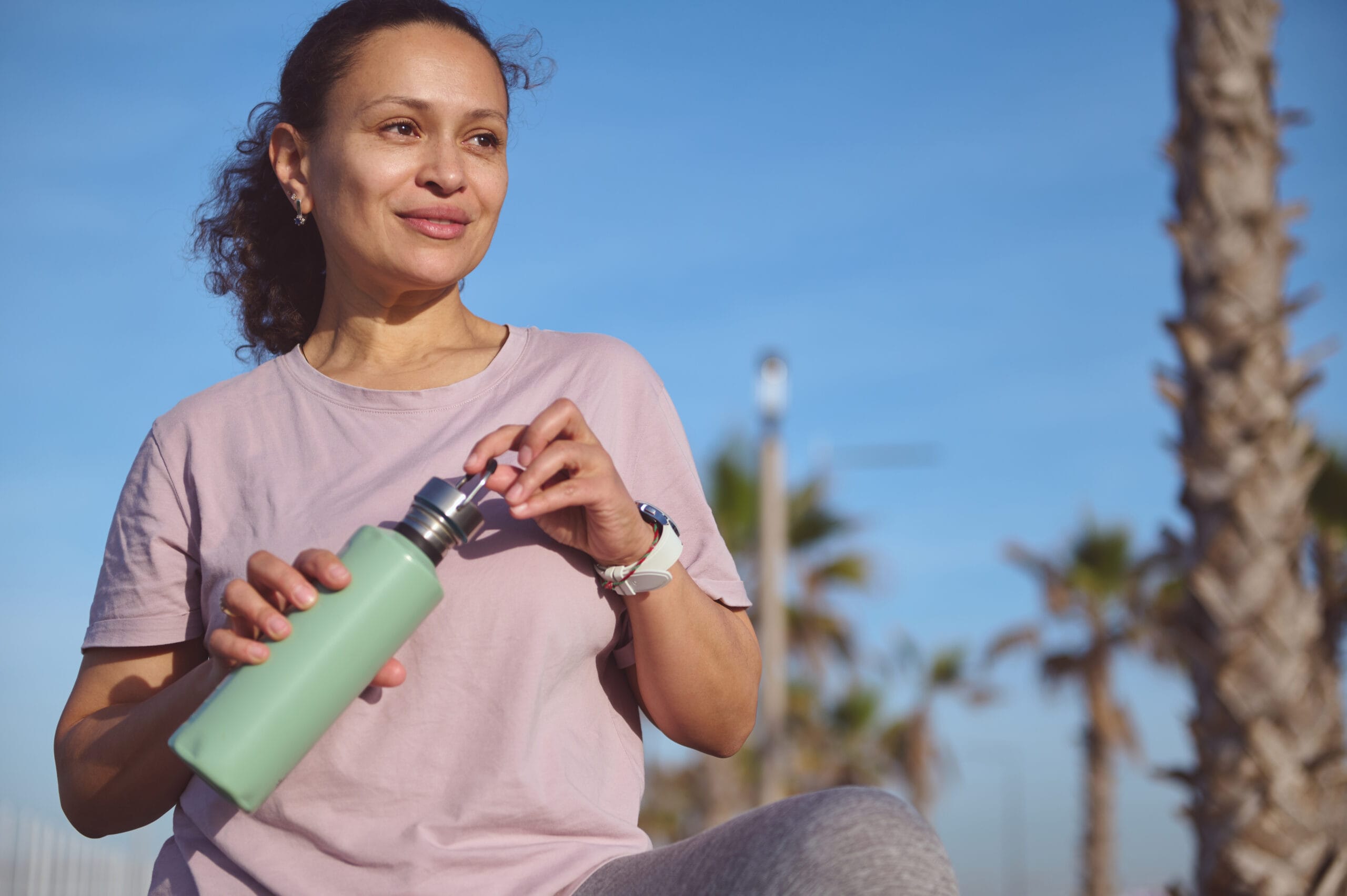 Authentic portrait of a multi ethnic woman, casually dressed, standing outside holding a green water bottle with a slight smiles on her face. There are palm trees and a blue sky behind her.