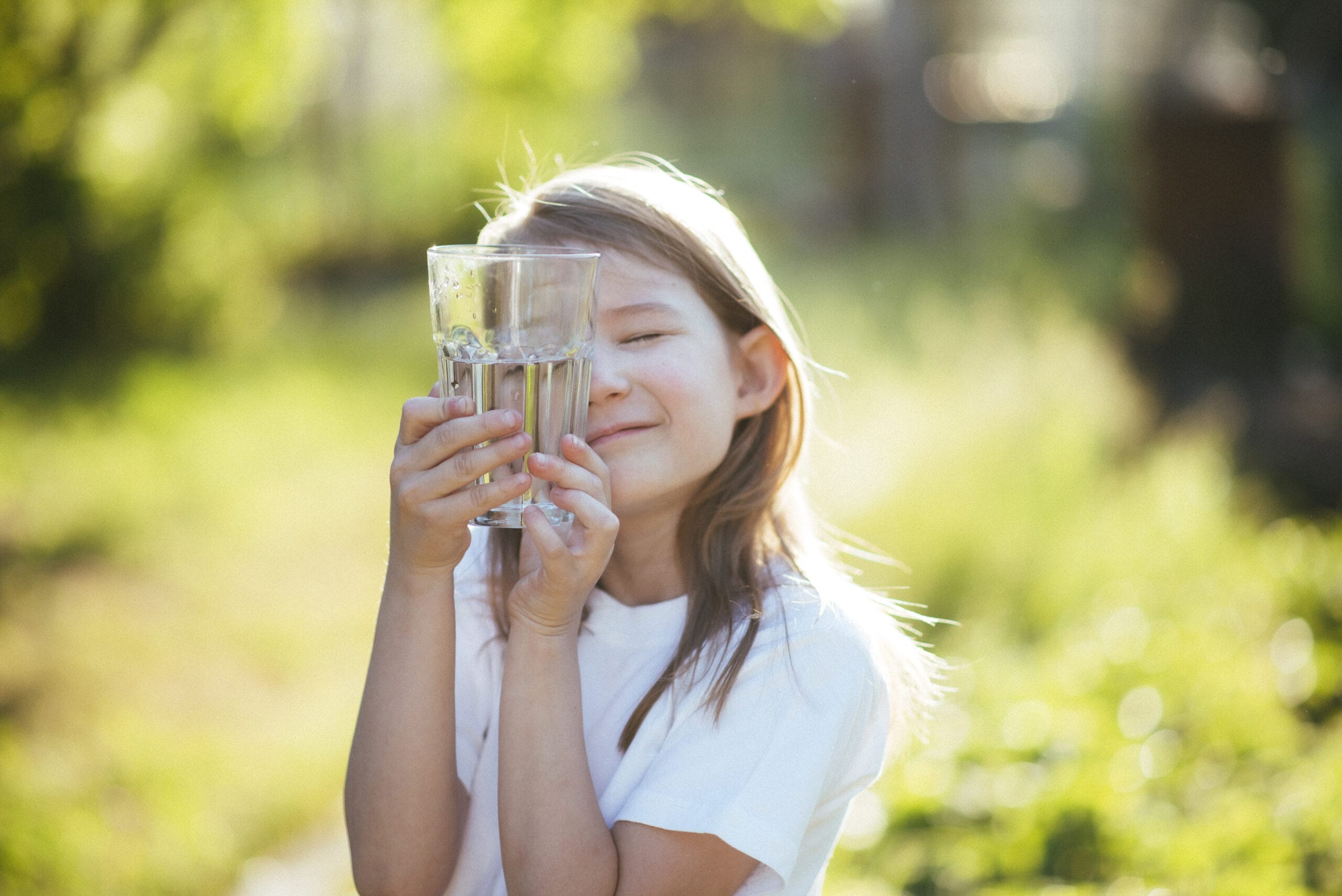 Portrait of a smiling little girl drinking water from a glass in nature on a sunny day.