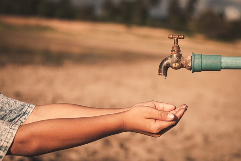 A rusty faucet drips water into a person's outstretched hands against a dry, barren landscape.