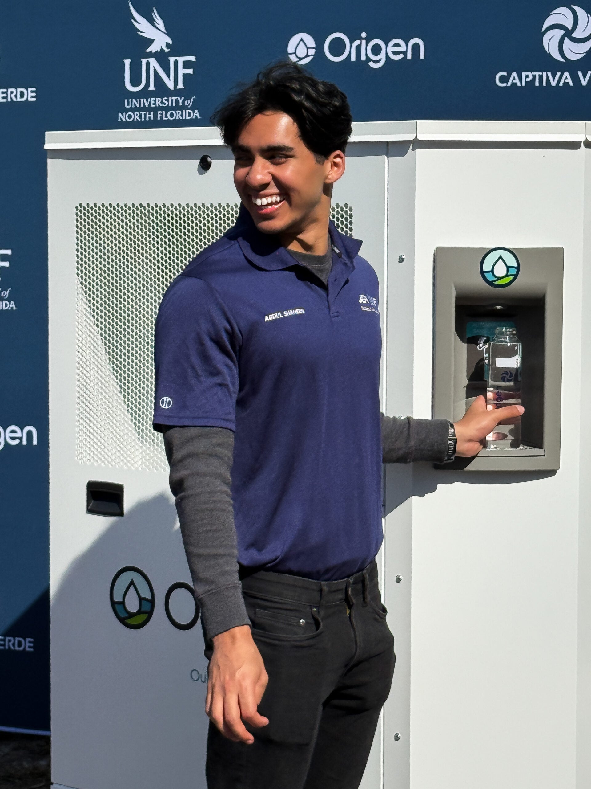 Smiling young man in a blue shirt stands next to a water dispenser, promoting sustainability at a university event.