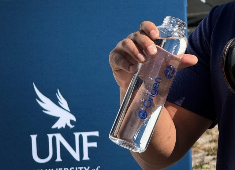 A hand holds a clear water bottle with the logo "Origen," set against a blue background featuring the UNF emblem.