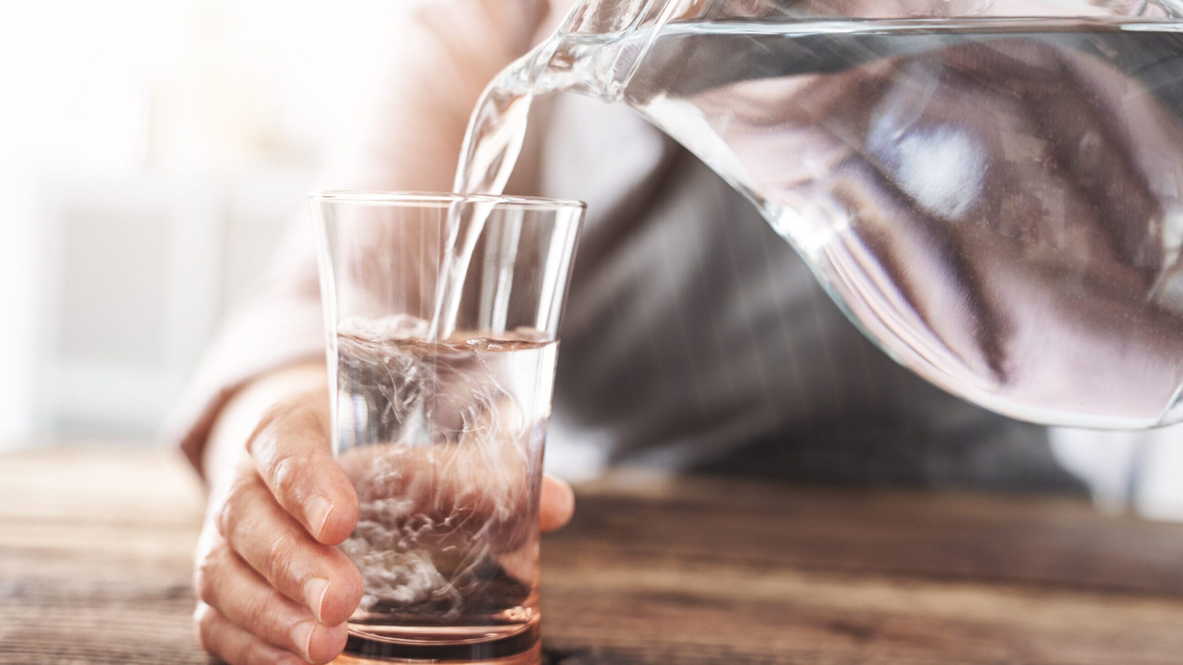 Clear water is being poured from a glass pitcher into a tall glass, with a hand gently holding the glass.