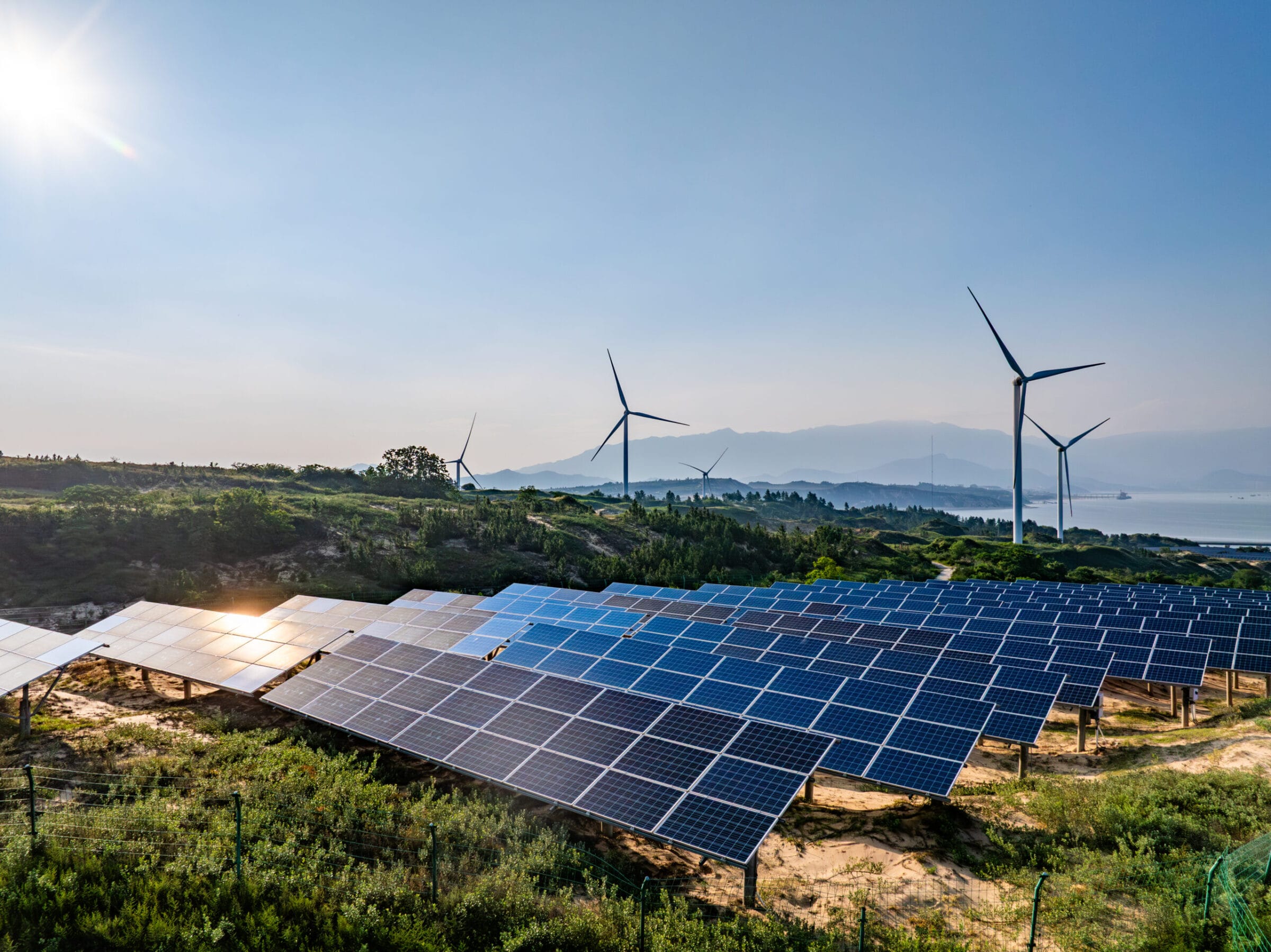 Solar panels in the foreground with wind turbines on a hillside under a clear blue sky and distant mountains.